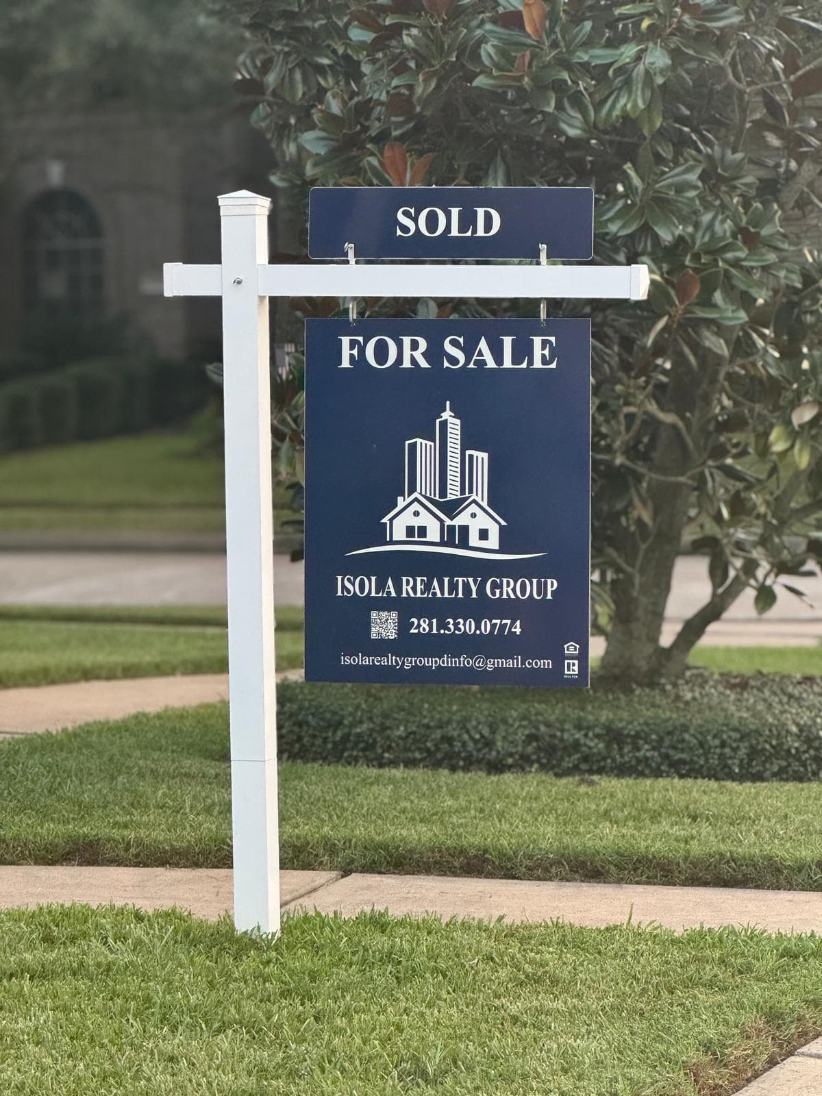 A navy Isola Realty Group for-sale sign with a sold rider, on a white post in a front lawn